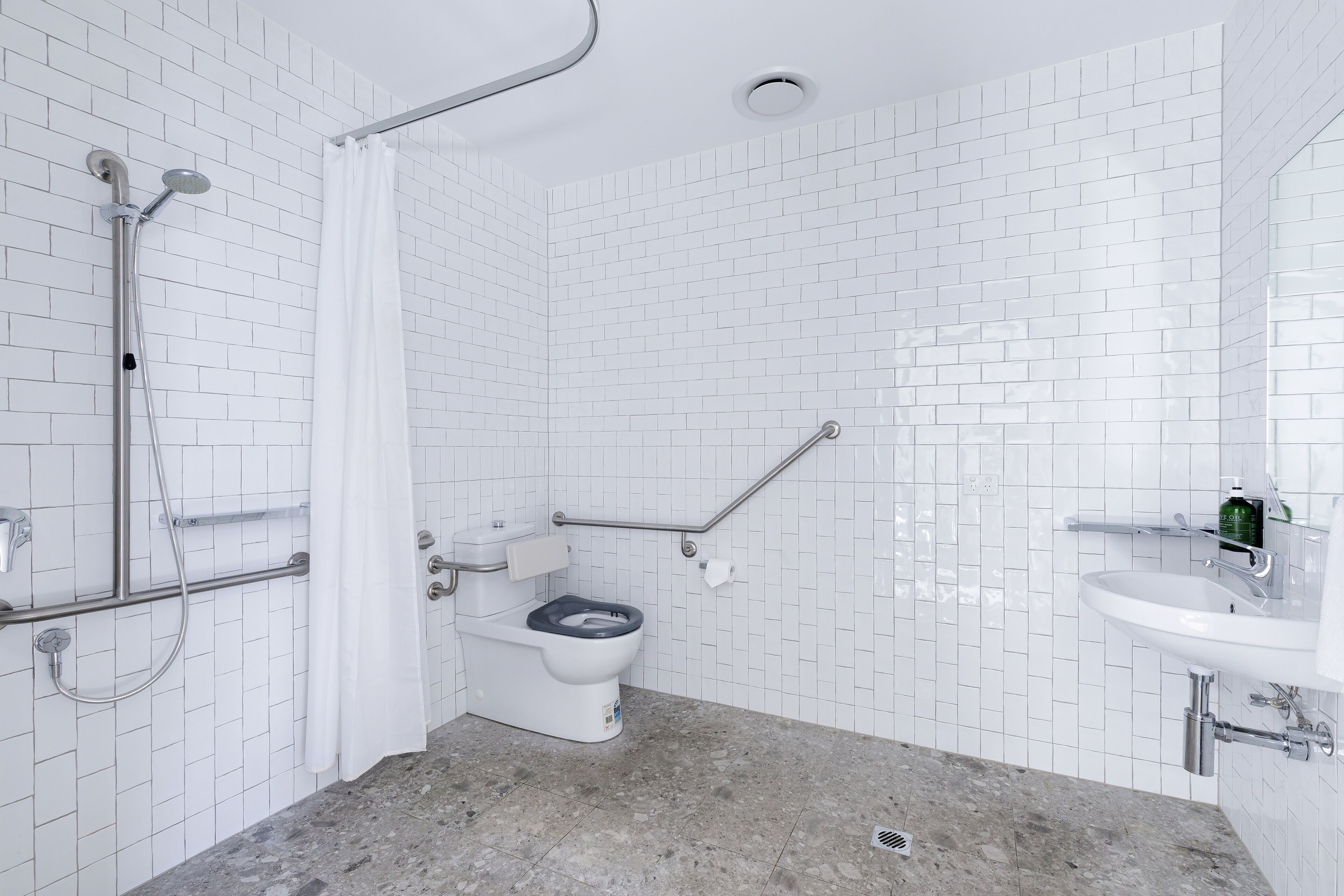 A modern bathroom with white subway tiles, a white toilet with a black seat, a shower with a handheld showerhead, a grab bar, and a sink with a green bottle on the counter.