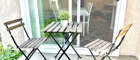 Une terrasse extérieure avec une table et deux chaises, un pot de fleurs et une porte vitrée donnant sur un espace intérieur.