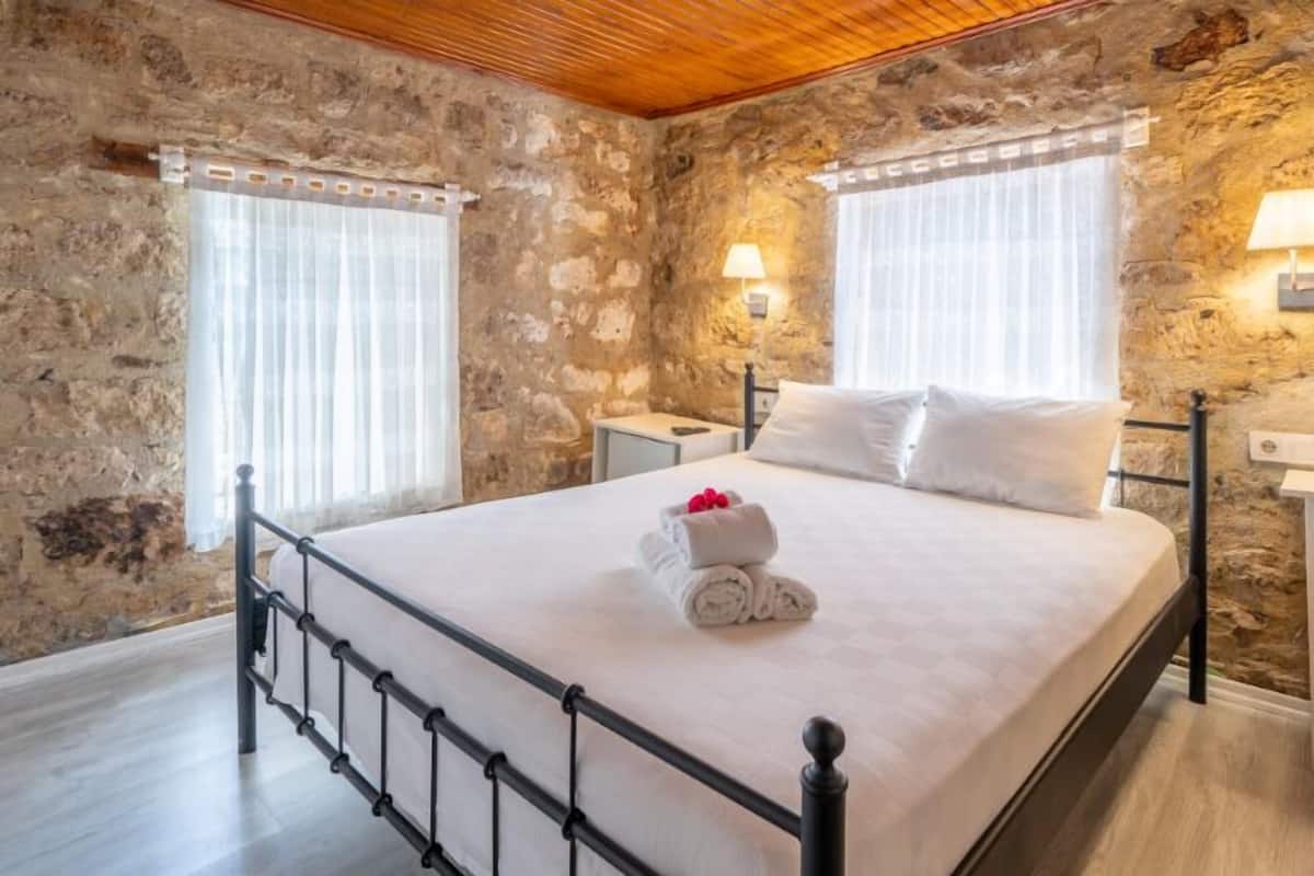 Un dormitorio con paredes de piedra, una cama con ropa blanca, dos mesitas de noche y un techo de madera.