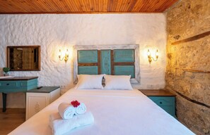 A bedroom with a stone wall, wooden ceiling, a bed with white linens, two nightstands, a dresser, and wall-mounted lights.