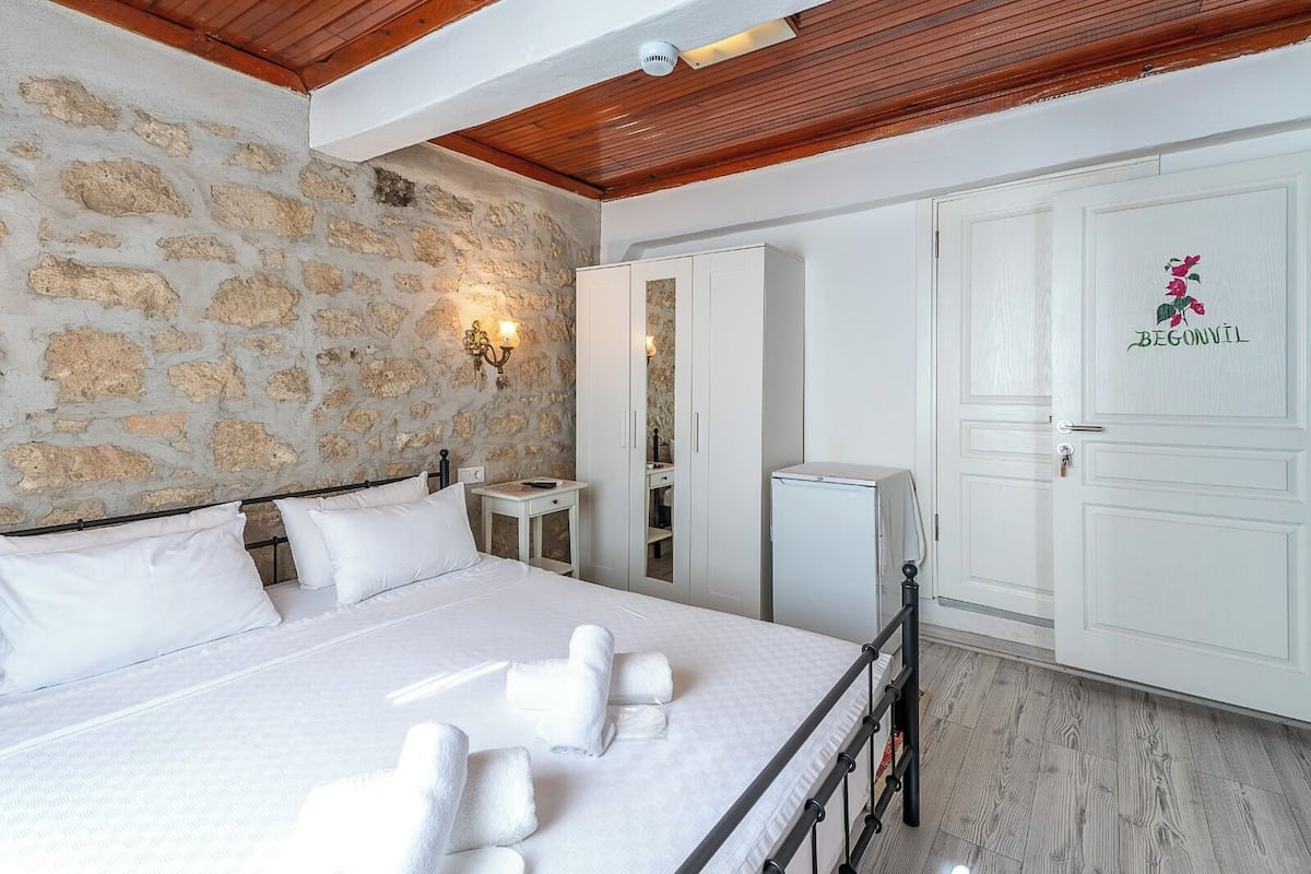 Un dormitorio con pared de piedra, una cama con ropa blanca, una mesita de noche pequeña, un armario y una puerta con placa.