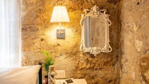 A rustic room with a stone wall, a wall-mounted lamp, a mirror, a bedside table with a book and a glass jar.