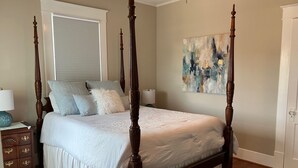 Ein Bett mit Baldachin, mit weißer Bettdecke und mehreren Kissen, ein Nachttisch aus Holz mit Schubladen, eine Lampe und ein Bild an der Wand.
