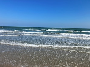 Uma praia com ondas, céu claro e barcos ao longe.
