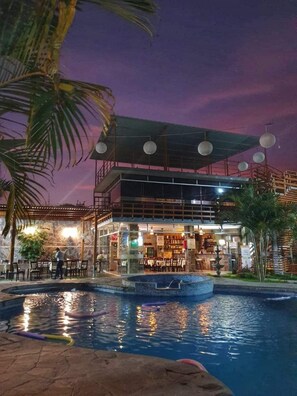 A pool area with a bar and seating, surrounded by palm trees and a building with a covered patio. - Gran Hotel Villa Del Sur (San Vicente de Cañete)