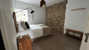 Ein Schlafzimmer mit Steinwand, einem Bett mit weißen Leinen, einer Holzbank und einem Schreibtisch mit Lampe.