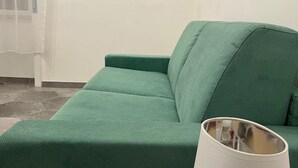 Vihreä sohva, jonka vieressä on valkoinen lamppu, seinällä on kaksi kehystettyä taideteosta ja ikkunassa on läpikuultavat verhot.