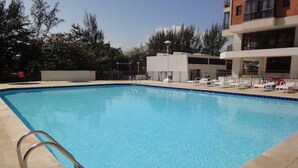 A swimming pool with a metal ladder, surrounded by lounge chairs and a building with a flat roof.