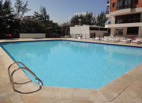 A swimming pool with a metal ladder, surrounded by lounge chairs and a building with a flat roof.