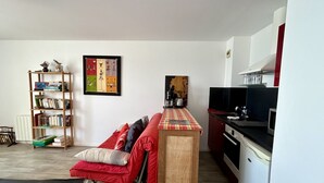 A compact living space with a sofa, a small kitchen area, a bookshelf, and a wall-mounted painting. - Les chouettes, apartment for 4 people (Le Crotoy)