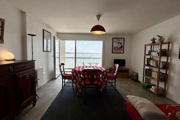 Sala de estar com mesa de jantar, cadeiras, sofá, estante de livros e vista para o mar.