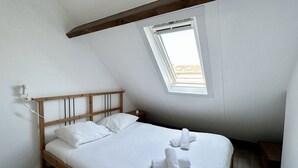 A bedroom with a sloped ceiling, a wooden bed frame, white bedding, a bedside table, and a window with a view. - Les chouettes, apartment for 4 people (Le Crotoy)