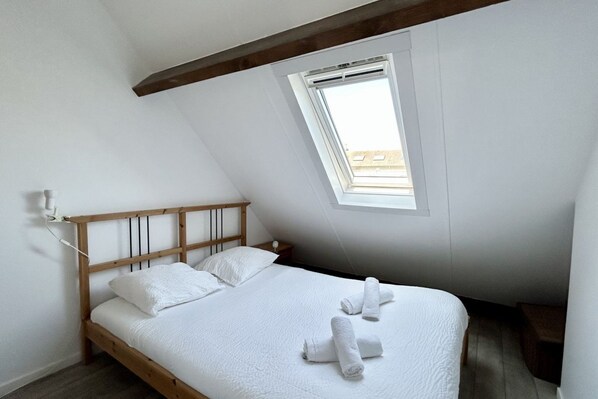 A bedroom with a sloped ceiling, a wooden bed frame, white bedding, a bedside table, and a window with a view. - Les chouettes, apartment for 4 people (Le Crotoy)
