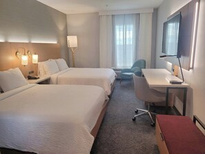 Ein Hotelzimmer mit zwei Betten, einem Schreibtisch, einem Sessel, einem Fernseher und einem Fenster mit Vorhängen.