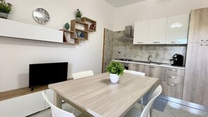 Une cuisine moderne avec des meubles blancs, une table à manger en bois et des chaises blanches. On y trouve un téléviseur à écran plat posé sur un meuble, une plante en pot sur la table et une petite étagère avec des livres et des objets décoratifs.