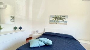 Une chambre à coucher avec un lit, une table de chevet, une lampe, un tableau représentant des palmiers et un miroir au mur.