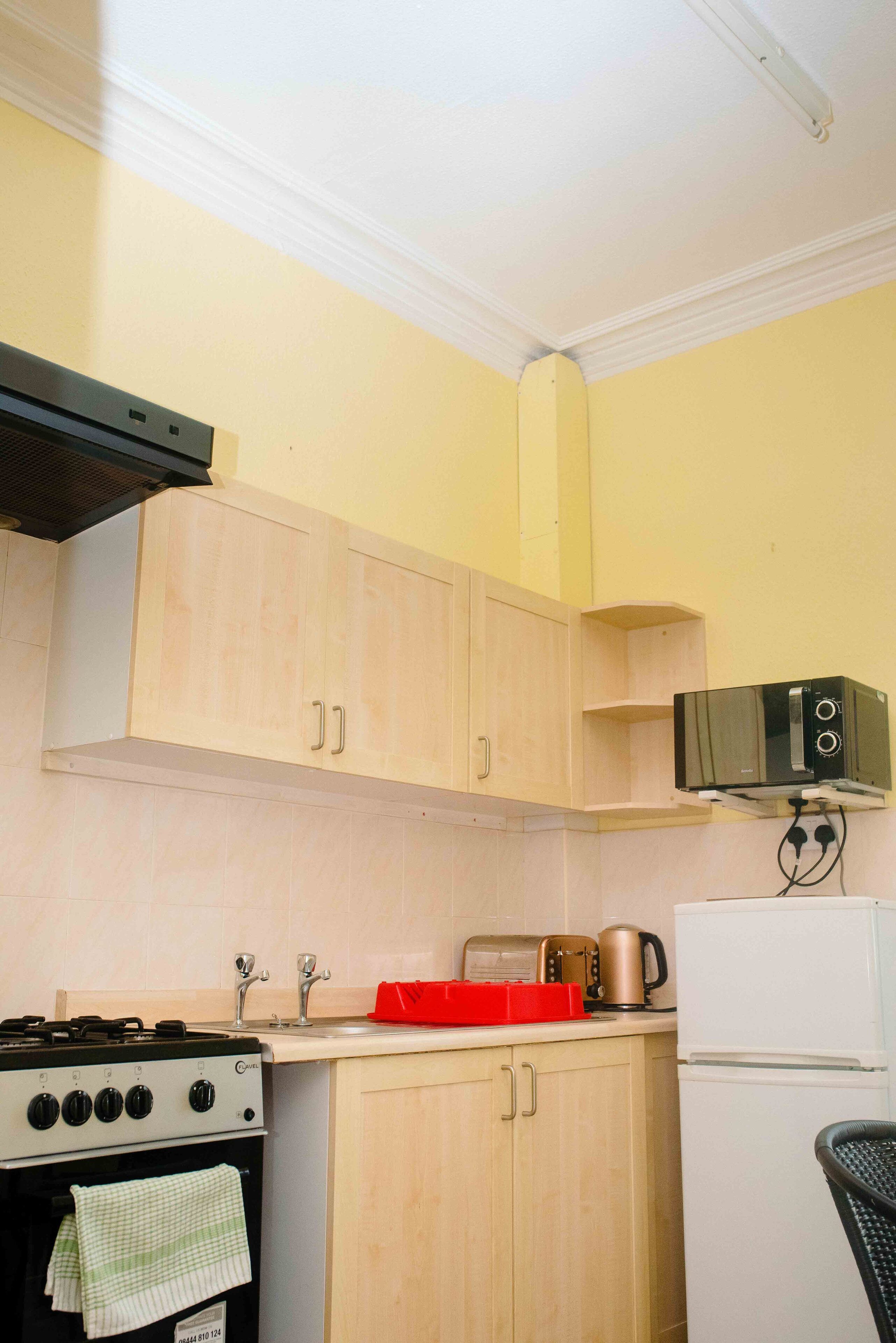 A compact kitchen with light wooden cabinets, a gas stove, a microwave, a kettle, and a refrigerator.
