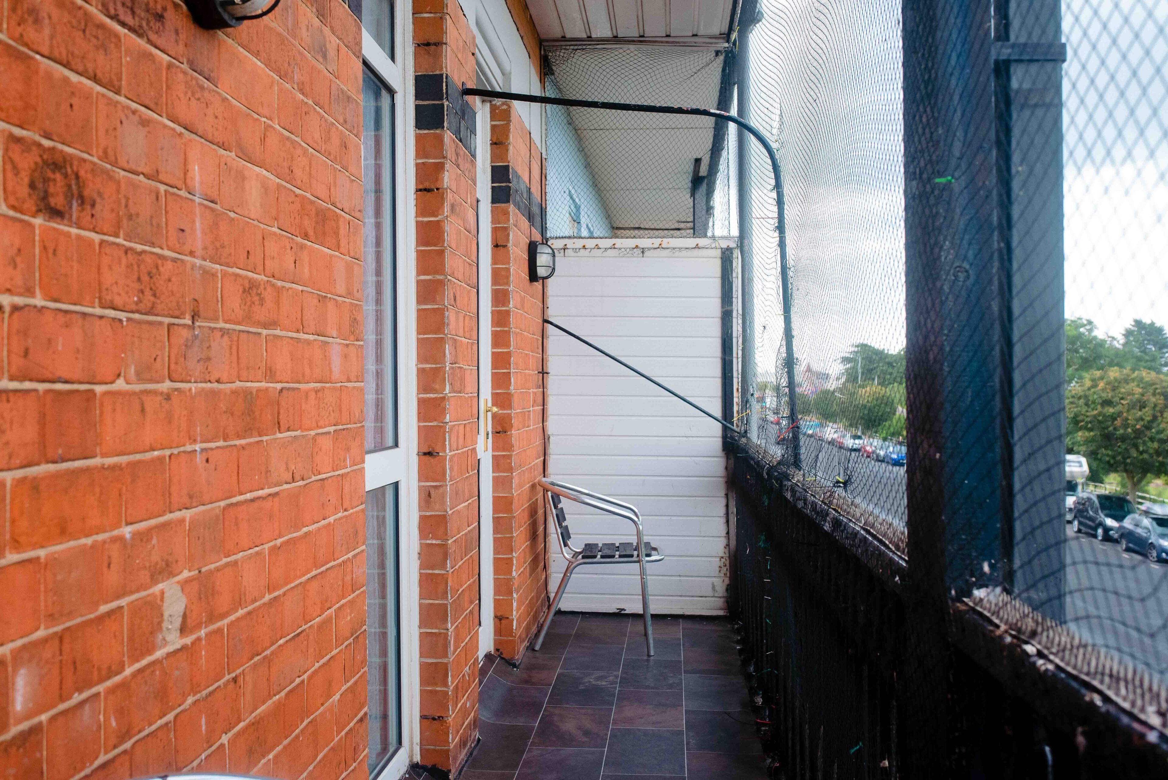A narrow outdoor corridor with a metal chair, a white door, and a chain-link fence.