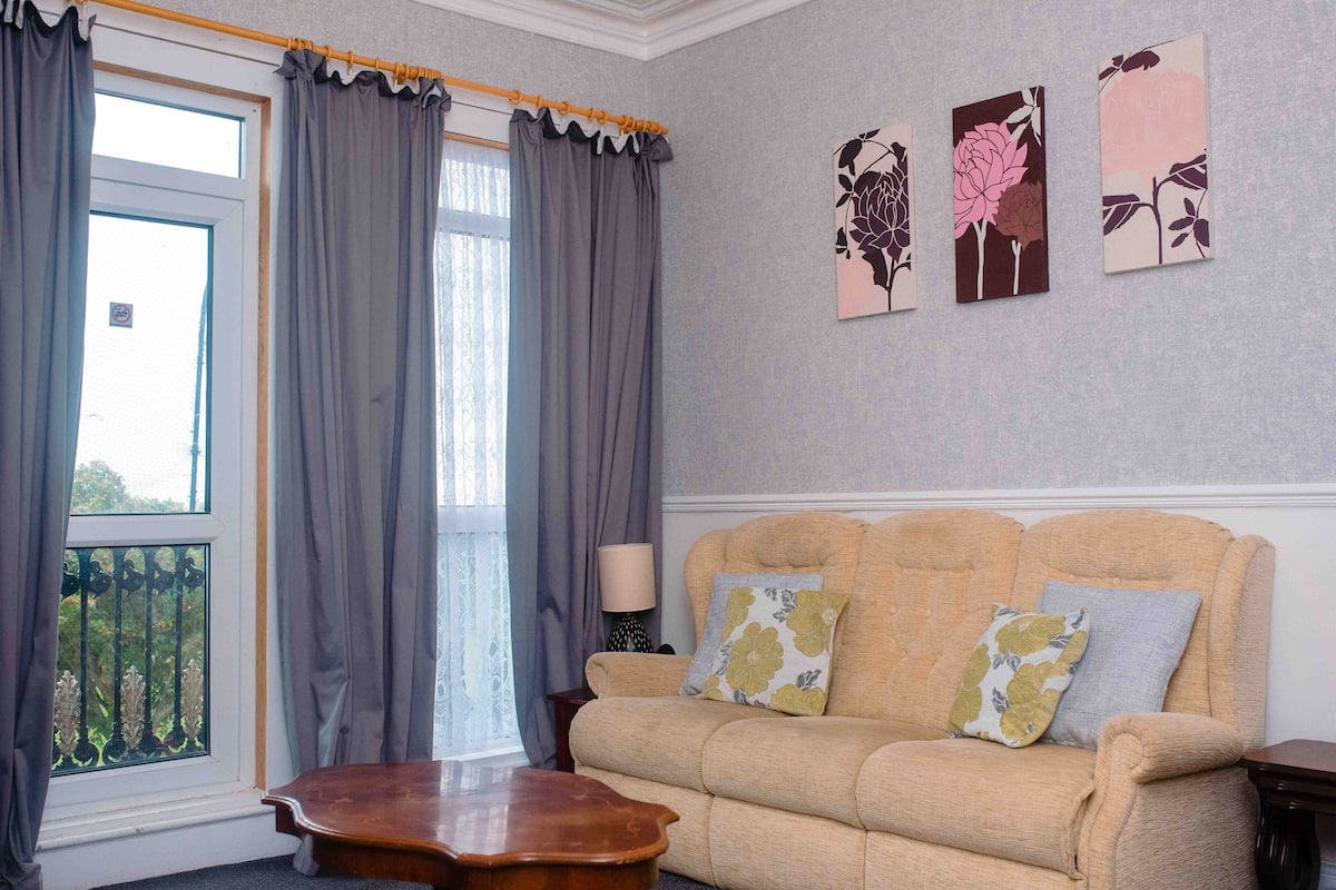 A living room with a beige sofa, two floral cushions, a wooden coffee table, and a window with grey curtains.