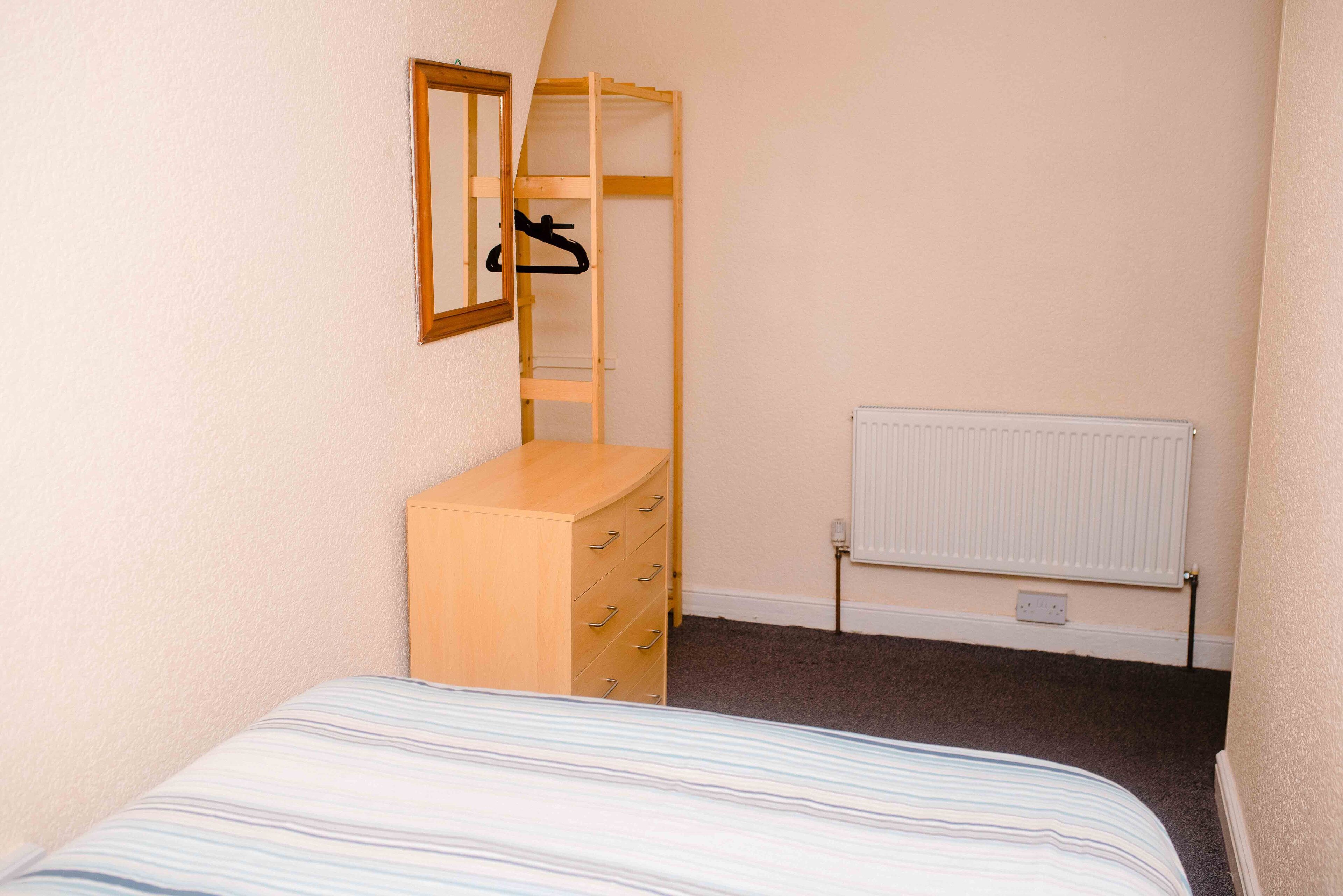 Ein kleines Schlafzimmer mit einem Bett, einer Holzkommode, einem Spiegel und einem Heizkörper.