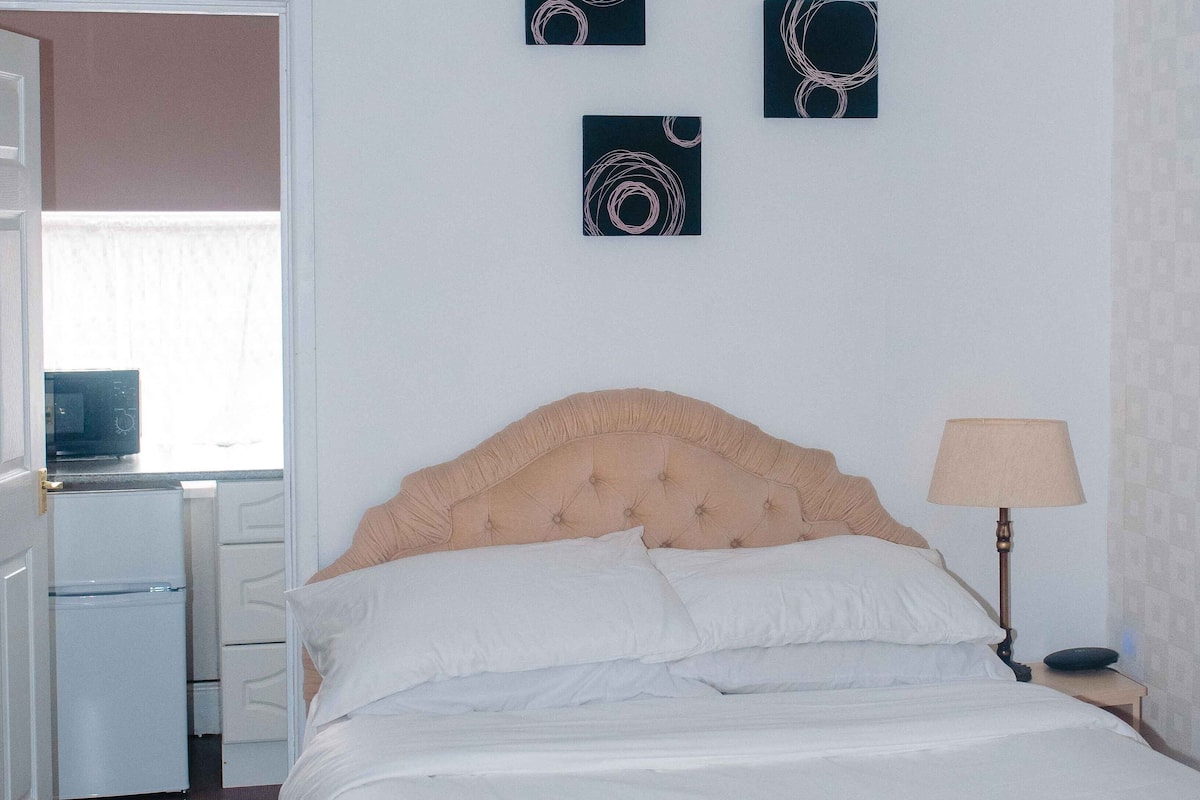 A bedroom with a bed, a nightstand with a lamp, a dresser, and a microwave. There are three wall art pieces above the bed.