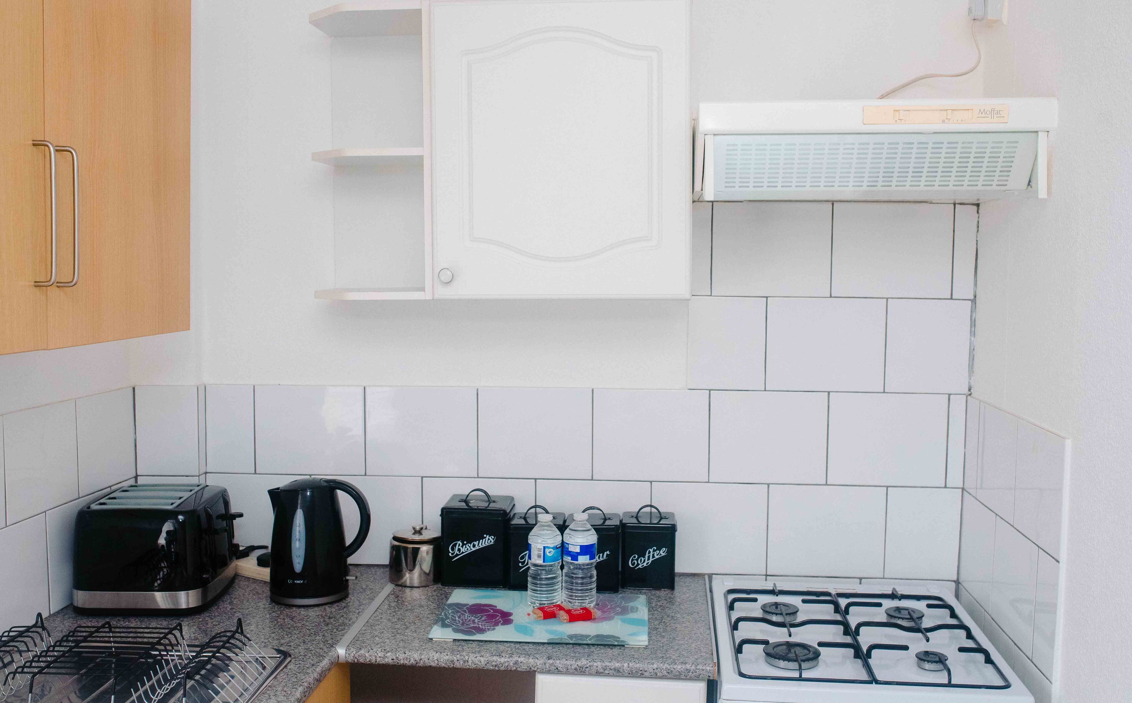 Una cucina con piano cottura a gas, un tostapane, un bollitore e una macchina per il caffè. Si vedono anche due bottiglie d'acqua e una scatola di biscotti.