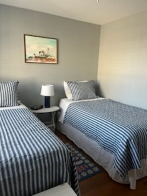 A room with two beds, a small table with a lamp, and a painting on the wall. - Cozy Cottage by the Sea (Kennebunk)