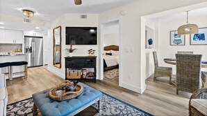 A modern kitchen with stainless steel appliances, a dining area with wicker chairs, and a living room with a blue sofa and a ceiling fan.