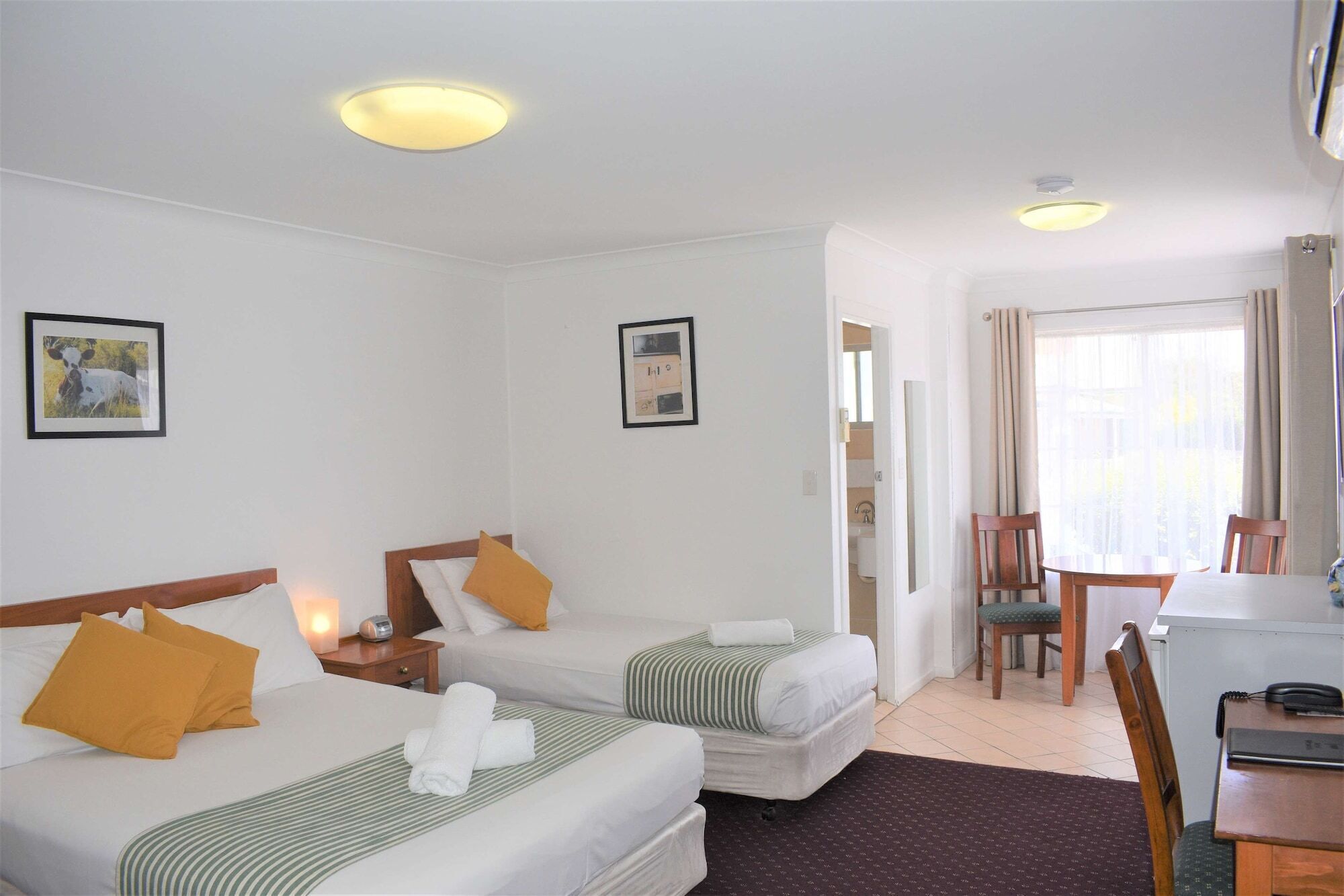A hotel room with two beds, a desk, and a chair. There are two framed pictures on the walls, a window with curtains, and a ceiling light.
