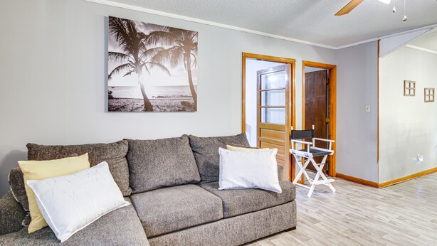 A living room with a grey sofa, white and yellow cushions, a director's chair, a large beach-themed painting, and a wooden door leading to another room. - Quiet & Cozy Rembert Home ~ 9 Mi to Air Force Base (Rembert)