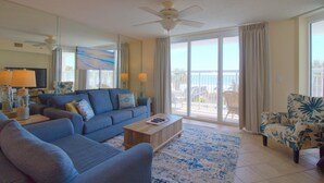 A living room with a blue sofa, a wooden coffee table, and a floral armchair. There is a ceiling fan, a large window with curtains, and a view of the outdoors. - Oceanfront 3BR/3BA Crescent Shores North Myrtle Beach (North Myrtle Beach)