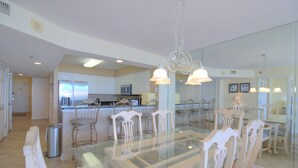 A spacious dining area with a glass-top table surrounded by white chairs, a kitchen in the background, and pendant lighting. - Oceanfront 3BR/3BA Crescent Shores North Myrtle Beach (North Myrtle Beach)