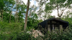 Una cabaña apartada rodeada de un bosque denso, con una terraza de madera y un amplio ventanal con vistas al follaje.