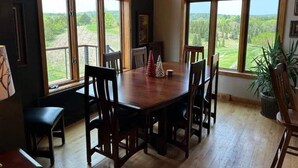Et spiseområde med et træbord og stole, store vinduer med udsigt til grøn vegetation, samt et træloft.