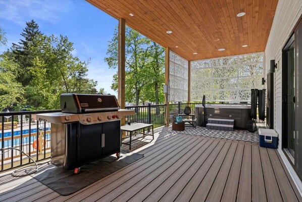 A covered outdoor deck with a grill, seating, and a hot tub.