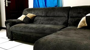 Un canapé de couleur sombre, agrémenté de deux coussins, se trouve dans une pièce dont le sol est recouvert de carreaux et où l’on aperçoit une porte fermée au fond.