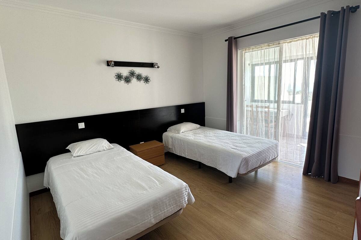 A hotel room with two single beds, a wooden floor, a black headboard, a small wooden nightstand, a wall-mounted shelf with decorative items, and a large window with curtains.
