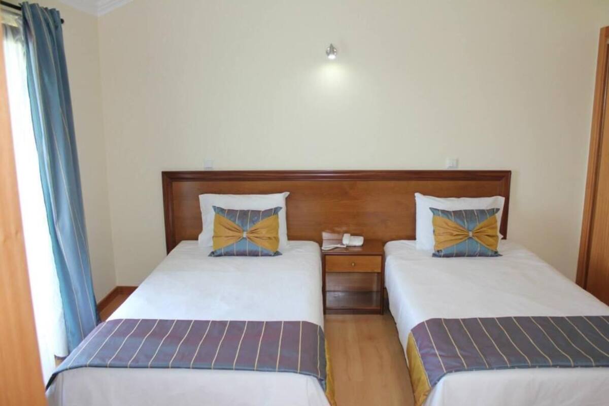 A hotel room with two single beds, each with a wooden headboard, a bedside table with a phone, and a curtain on the left side.