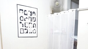 A small, neatly arranged room with a grey sofa, a wooden chair, a bed with patterned bedding, a wall-mounted fan, and a shower curtain. - Bed Space Hostel  (Bangkok)