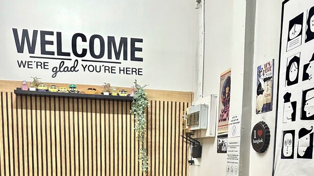 A cozy room with a wooden slat wall, a welcome sign, a small table with chairs, and a grey sofa. - Bed Space Hostel  (Bangkok)