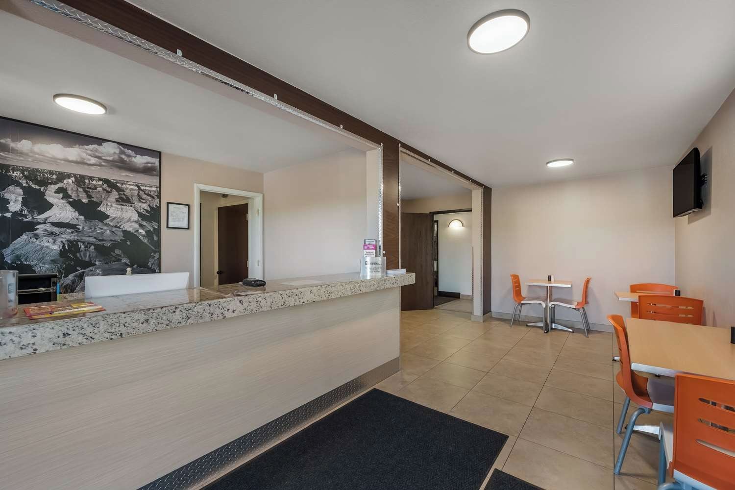A hotel lobby with a reception desk, a large painting, a dining area with orange chairs, and a flat-screen TV mounted on the wall.