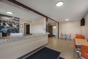 Un hall d’hôtel comprenant un comptoir d’accueil, un grand tableau, un espace repas avec des chaises orange et un téléviseur à écran plat fixé au mur.