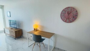 A modern room with a wooden desk, a black chair, a small table with a lamp, a TV on a wooden cabinet, and a round wall clock. - French Ocean View , Beach and Pool (Cul-de-Sac)