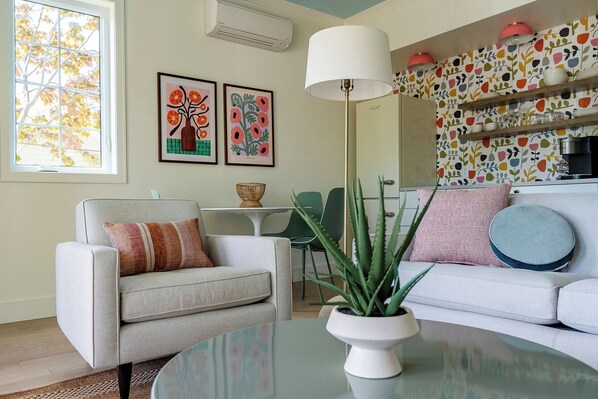 Un salon comprenant un canapé, un fauteuil et une table basse agrémentée d’une plante. On y trouve une fenêtre donnant sur des arbres, un mur orné de tableaux encadrés et un coin cuisine avec des étagères.
