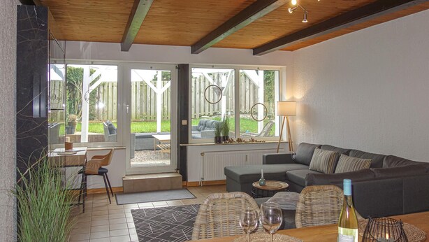 Een gezellige woonkamer met een bank, een eettafel met wijnglazen en uitzicht op een tuin door grote ramen.