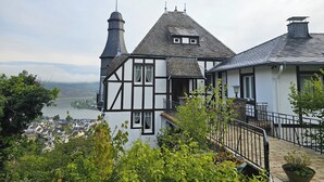 Una casa tradizionale a graticcio con una torre imponente, vista su un fiume e una città sullo sfondo.