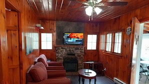 A cozy cabin interior with a stone fireplace, wooden walls, a ceiling fan, and a television mounted above it.