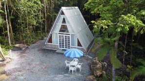 Een klein huis met schuin dak en een groot raam, omgeven door bomen en een grindpad.
