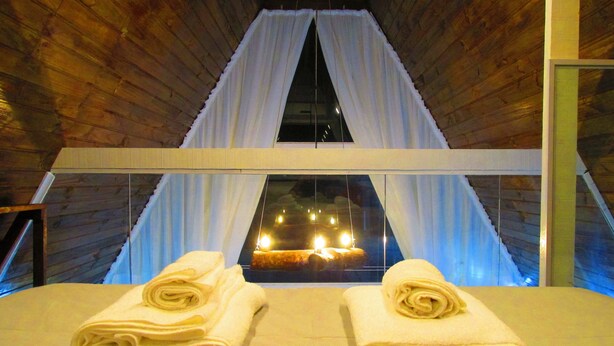 Een kamer met een bed, twee opgerolde handdoeken en uitzicht op een verlicht interieur door een raam met witte gordijnen.