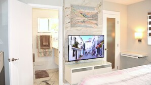Une chambre d’hôtel avec un téléviseur fixé à un mur de marbre, un lit recouvert d’une courtepointe blanche et rose, et une petite étagère avec une lampe.